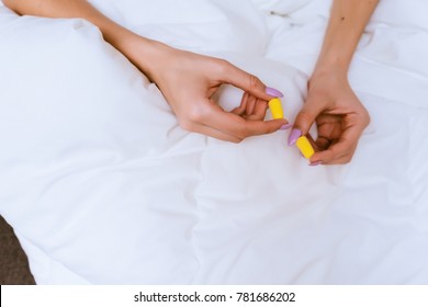 the girl sits on a bed under a white blanket, in hands yellow earplugs against street noise