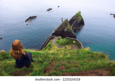 Girl sits on a cliff over the ocean and the Dunquin Pier situated on the west coast of the Dingle Peninsula in Ireland - Shutterstock ID 1231024318