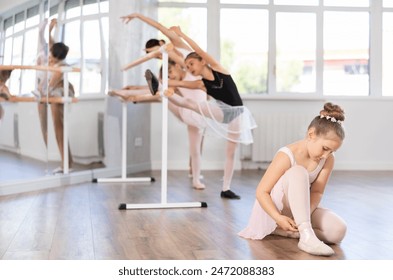 Girl sits on floor and adjusts her shoes, ties ribbons tightly around her shin and legs in ballet studio. Child pupil adjust pointe shoes before start of training - Powered by Shutterstock