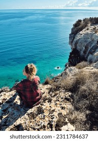 The girl sits on a high stone and looks at a beautiful view of the blue sea and sky. Coastline. Cagliari. Sardinia, Italy. Vertical image - Shutterstock ID 2311784637