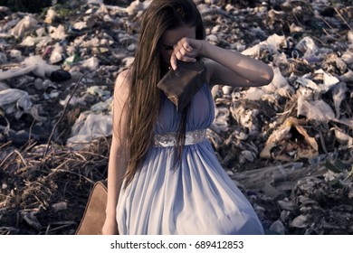 The Girl Sits On A Suitcase Among A Garbage Dump With An Empty Purse And Cries