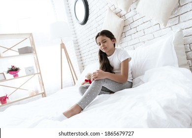 Girl Sits On A White Bed And Holds Her Hand Over A Sore Knee. Little Girl Injured A Knee Sitting On A Bed With A Sad Face. 