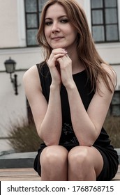 A girl sits on a wooden bench in the street and looks at the camera. a Girl sits on a wooden surface and looks at the camera. a Girl looks at the camera. portrait of a girl in a black dress