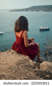 
A girl sits over a cliff.Woman on a rock.Resting on the sea.Mediterranean sea.Travel agency.Lady in red.Portrait of a girl at sunrise.Sea tourism and vacation.Woman looks at the sea.Calmness.Pleasure - Shutterstock ID 2255053011