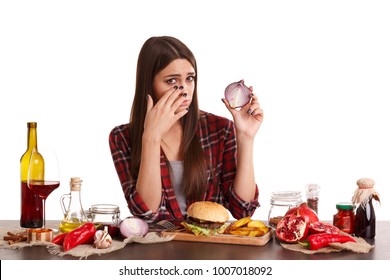 A girl sits at a table with food and holds a half of onions and cries from it. Isolated on white.