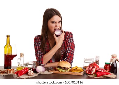 A girl, sits at a table with food and holds a half of onions and bites it. Isolated on white.