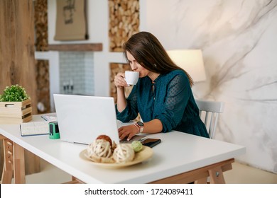 A Girl Sits At A Table In The Office Filling A Contract Or Financial Report With A Cup Of Coffee. Types On A Laptop. There Are Documents On The Table. Image With Selective Focus