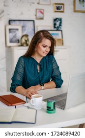 A Girl Sits At A Table In The Office Filling A Contract Or Financial Report With A Cup Of Coffee. Types On A Laptop. There Are Documents On The Table. Image With Selective Focus