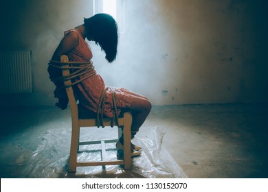 Girl is sitting alone. Her hands and legs are tied with ropes to the chair. Girl is looking down. She has lost consciousness. She is sitting in front of small window.