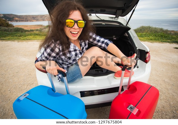 Girl sitting in back of car smiling and showing\
thumbs up. Young laughing woman sitting in the open trunk of a car.\
Summer road trip