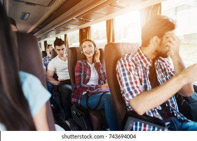 The girl is sitting on the bus in headphones and listening to music. She smiles and enjoys music. Next to the bus are other people who also went on a trip. - Powered by Shutterstock