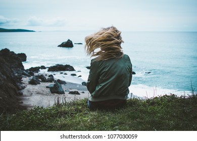 Girl Sitting On A Cliff Overlooking The Ocean With Her Hair Blowing