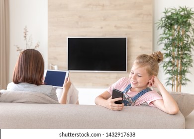 Girl Sitting On Couch And Watching Something Interesting In Mobile Phone While Mother Watching Tv. Surprise Concept