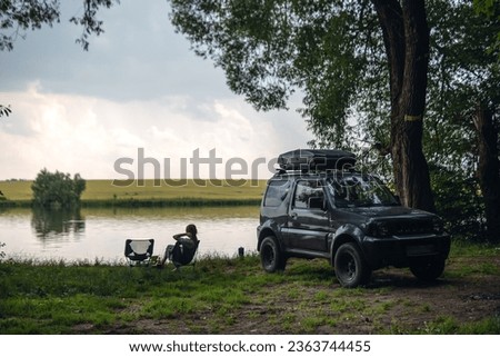 The girl is sitting on a folding chair. Picnic. off-road vehicle on the shore of the lake, forest in the middle of wilderness. Without recognition. The concept of extreme recreation on an SUV vehicle