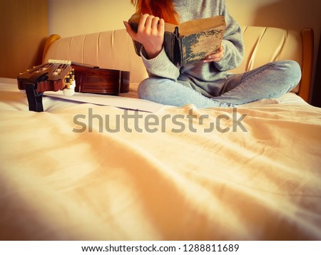 Girl sitting on her bed and reading an old book near her is put a guitar view from down up