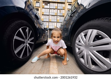 A Girl Is Sitting On The Road Markings In The Parking Lot Between Two Cars On A Warm Summer Day. A Multi-storey House In The Background Behind A Child