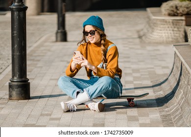 Girl sitting on skateboard and use mobile phone. Outdoors, urban lifestyle. cute skater girl sitting on skate board checking smart phone listening to music using internet and takes a photo. - Powered by Shutterstock