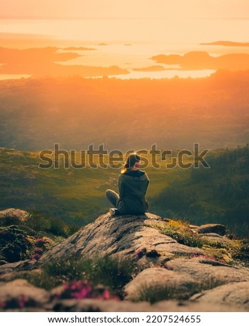 A girl sitting on the top of the mounting and enjoying yellow sunrise above sea. Hiking woman in khaki jacket relaxing on the cliff looking at a beautiful sunlit landscape. Green valley in sunlight