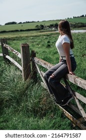 The girl is sitting on a wooden fence in the field. The girl sits on the fence and looks at the green fields.	