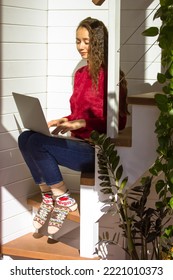 Girl Sitting On Wooden Staircase Steps Indoor Working On Laptop At Fall Winter Season. Smiling Young Woman Working Remotely, Studying, Looking For Information In Internet. Stay At Home. Vertical Photo