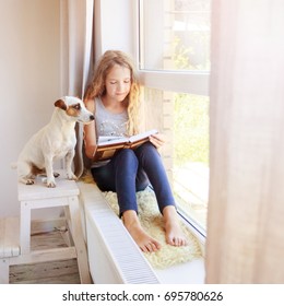 Girl Sitting At Window At Read. Child Reading Book At Home. 