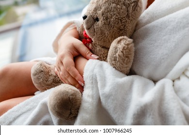 The girl sitting at the window in a white robe with a Teddy bear with a red bow in his hands. Closeup of woman's hand with a stuffed animal in his hands at the window.