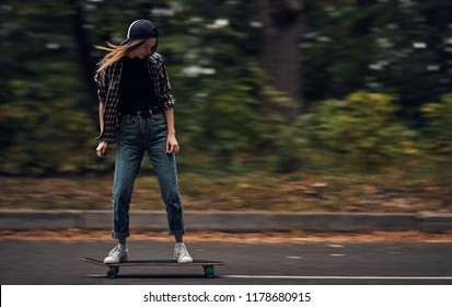 A girl skateboarder in a cap and checkered shirt rides a skateboard on the road in the forest. - Powered by Shutterstock