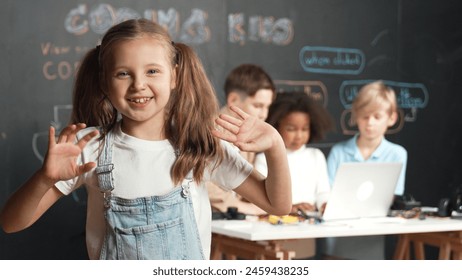 Girl smile and wave to camera while friend working or learning engineering code or prompt in STEM technology classroom. Student looking at camera and greeting while children using laptop. Erudition. - Powered by Shutterstock