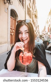 Girl, Smile Young Woman In Leather Jacket With Sunglasses And Brown Hair In A Cafe On The Street, Rome, Italy. Vacation In Italy, Europe. Portrait Of A Beautiful Woman Drinking Cocktail Aperol Spritz