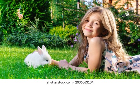 The Girl Smiles Cutely. A Child Plays With A White Fluffy Bunny In The Garden On A Green Lawn In Summer. Decorative Dwarf Rabbit Eating Grass On The Lawn Outdoors. Positive Emotions In The Child.