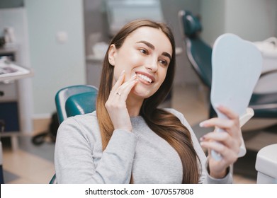 The Girl Smiles In Dentistry. Patient Dentist Racing Model At The Dental Clinic. White Well-groomed Teeth After Taking A Doctor. Tooth Whitening And Tooth Enamel. In The Hands Of The Mirror