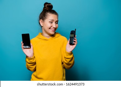 Girl Smiles At An Old Cell Phone, Which She Holds In Her Hand, Holding A Modern Smartphone In Her Second Hand