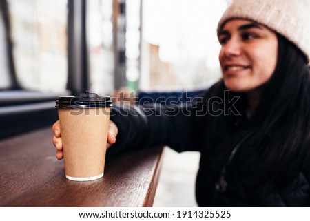 Girl smiling, holds paper cup of coffee in hand near cafe. Blurred background