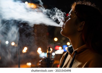 The girl smokes an electronic cigarette against the backdrop of the night city. Woman enjoying vape, nicotine steam vaporizer. Bad habit.