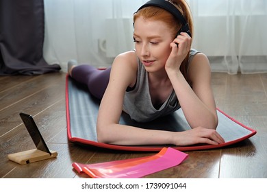 The girl in sports uniform and headphones lies on stomach on the mat and looks into the phone, supersing head with her hand