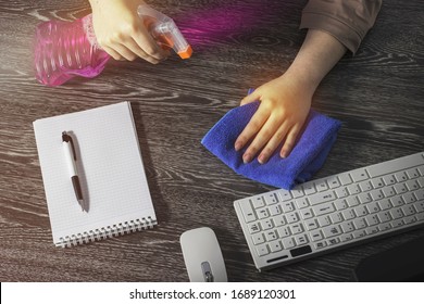 The girl sprays cleaning agent on the table and wipes it with a blue cloth. Keyboard, mouse and Notepad with pen on the office Desk. The concept of cleaning
