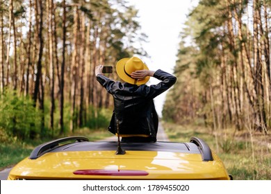 Girl Standing at Car Sunroof Enjoying Road Trip. Woman in Leather Jacket on Summer Getaway. Back View of Beauty Making Selfie Leaning out Yellow Auto Roof with Forest on Background