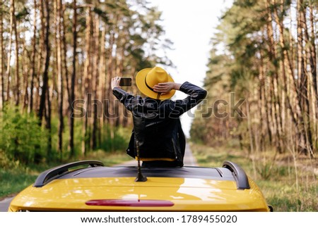 Girl Standing at Car Sunroof Enjoying Road Trip. Woman in Leather Jacket on Summer Getaway. Back View of Beauty Making Selfie Leaning out Yellow Auto Roof with Forest on Background