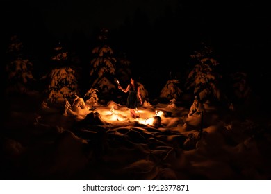 Girl Standing In Dark Deeply Snow-covered Forest She's Got Long Blond Hair And Is Wearing A Black Dress. She's Holding A Torch That Casts A Warm Cozy Light. All Of That Creates A Fairy Tale Atmosphere