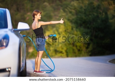 Girl standing next to electric car holding a charging cable and hitchhiking.