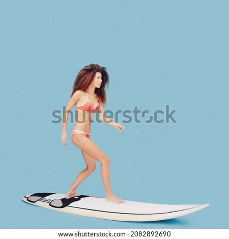 Girl standing on surfboard keeping balance ready to meet the wave of summer adventure, freshness, hurry to see the collections of new bikini over blue studio background. Side view.