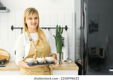 The girl stands in a bright kitchen and holds cupcakes with white cream and blueberries on a chocolate tray. Concept background, kitchen, confectioner, breakfast - Powered by Shutterstock