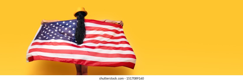 girl stands with her back and holds the American flag over yellow background, girl in fashionable hat with USA flag, panoramic mock up image