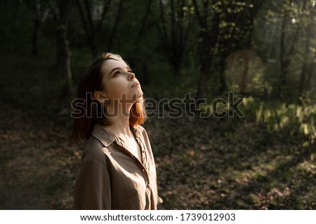 a girl stands in the middle of the forest and looks up at the rays of the sun envelop her face