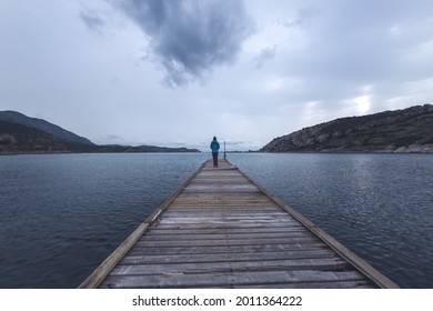 A girl stands on the pier and looks at the sea, a woman stands in the middle of the pier, a trip to picturesque places, a girl against the background of a lake, mountains and a cloudy sky, cold season