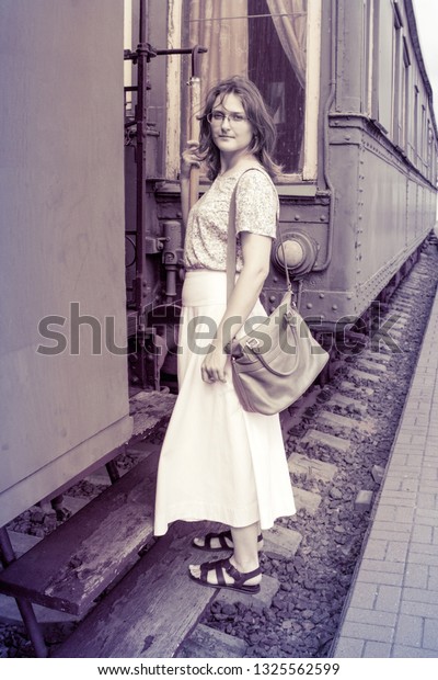 A girl stands on the steps of the cars\
train. Historical monochrome photo of a woman on the footboard of\
the car. The vintage train is at the\
station