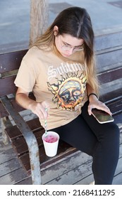 Girl Stirring Her Strawberry Slushie While Resting On A City Bench.