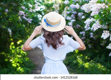 Girl in a straw hat with a blue ribbon on a spring afternoon. Back view. Trendy casual summer or spring outfit. Woman in a boatman's straw hat. concept of female spring fashion. Flowering lilac bushes - Powered by Shutterstock