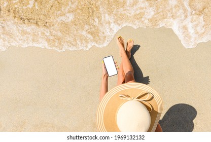 A Girl In A Straw Hat Sits On The Beach With A Phone In Her Hand. View From Above. Phone With Blank White Display For Your Text