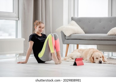 Girl Stretch Her Toes During Online Workout Lesson With Tablet And Looking At The Camera. Golden Retriever Dog Sleeping Close To Owner On The Floor